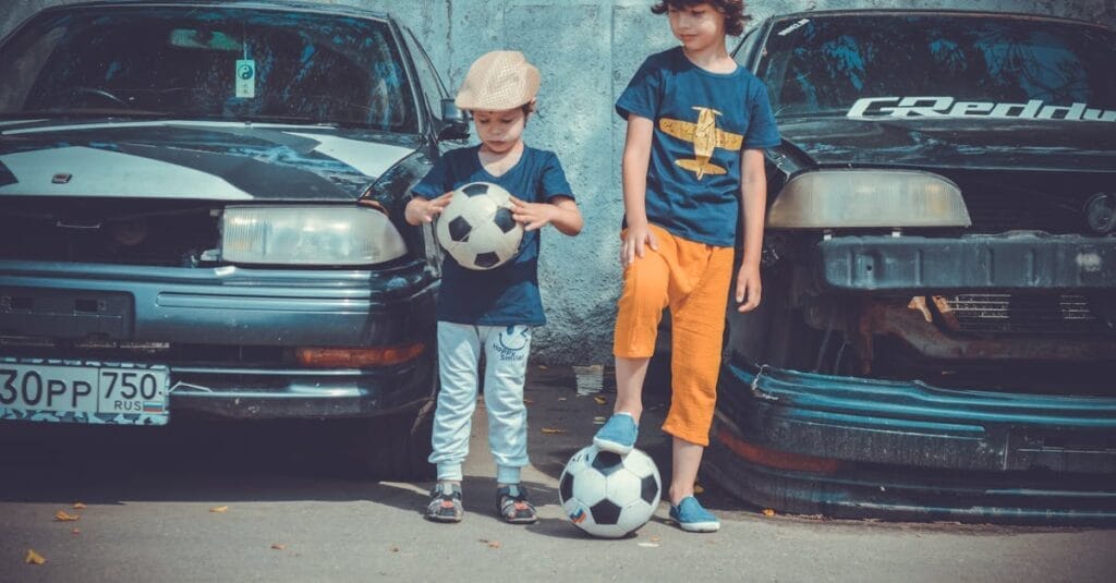 Two children in casual attire play soccer near vintage cars on a sunny day.