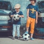 Two children in casual attire play soccer near vintage cars on a sunny day.