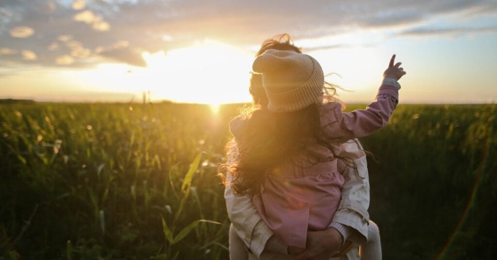 A mother and daughter embrace and point at the sunset in a grassy field.