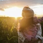 A mother and daughter embrace and point at the sunset in a grassy field.