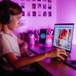 Child focused on laptop wearing headphones at desk with purple lighting. Modern tech scene.