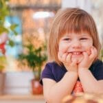 A happy young girl smiling indoors surrounded by plants and a pastry, with natural light streaming in.