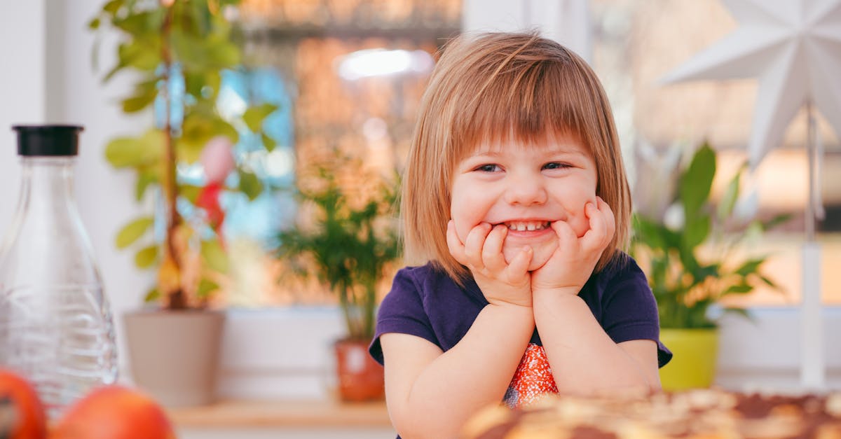 A happy young girl smiling indoors surrounded by plants and a pastry, with natural light streaming in.