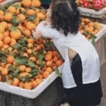 A child in traditional clothing leans over a market stall of fresh oranges.