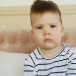 A young child sitting on a couch indoors, displaying a sad expression.