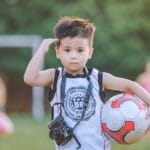 A young boy holding a soccer ball and camera stands confidently on a sunny day.