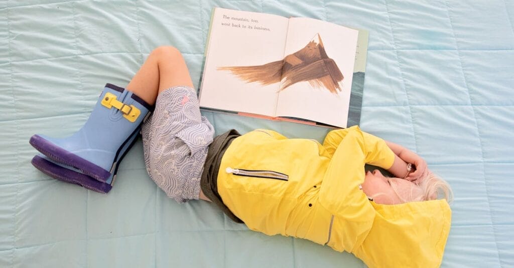 A child in a yellow raincoat and boots relaxes with a book on a light blue quilted bedspread.