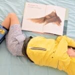 A child in a yellow raincoat and boots relaxes with a book on a light blue quilted bedspread.