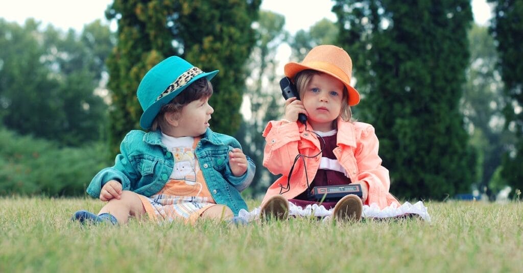 Two toddlers in colorful outfits enjoying playtime with walkie talkies in a sunny park setting.
