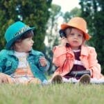 Two toddlers in colorful outfits enjoying playtime with walkie talkies in a sunny park setting.