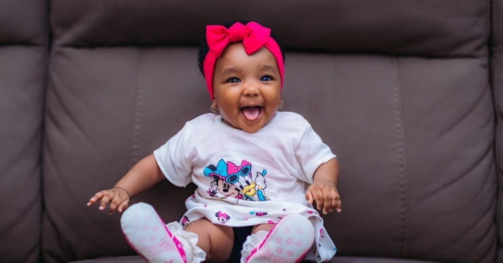 Cute baby with pink headband smiling joyfully on a sofa.