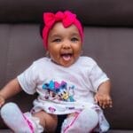 Cute baby with pink headband smiling joyfully on a sofa.