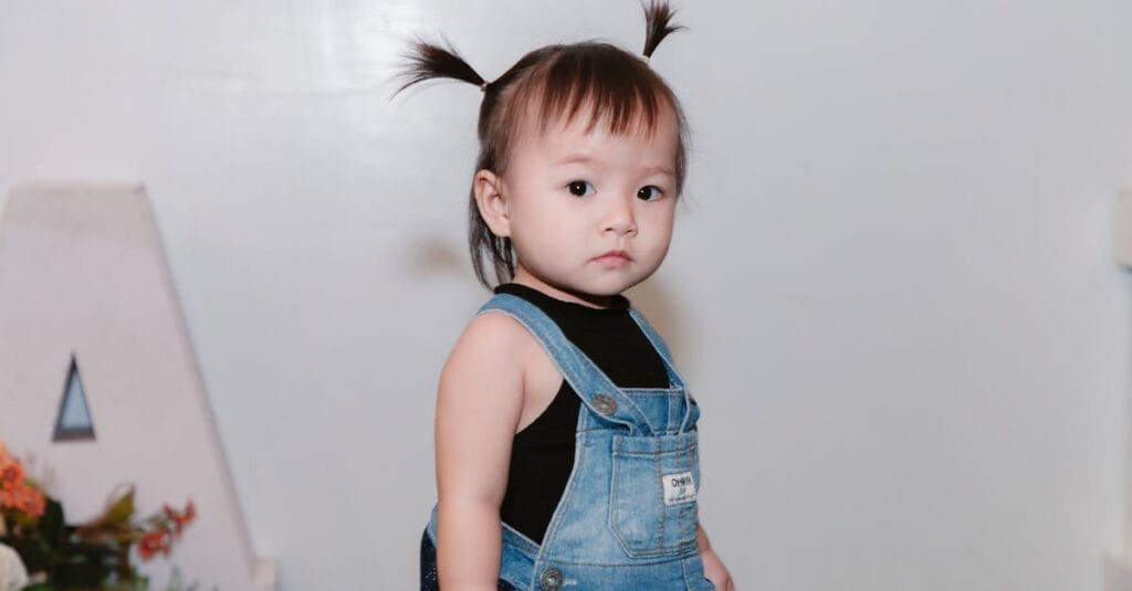 Adorable toddler with pigtails wearing a denim dress indoors.