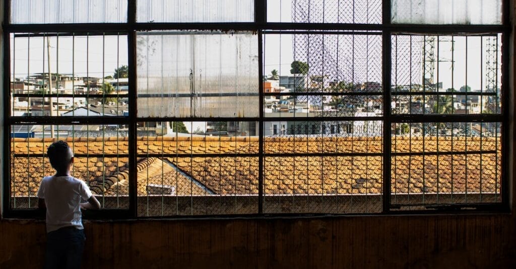 A child gazes outside through a large window, revealing an urban landscape with buildings in the background.