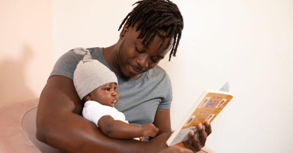 A father with dreadlocks reads a book to his baby, creating a bonding moment.