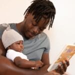 A father with dreadlocks reads a book to his baby, creating a bonding moment.