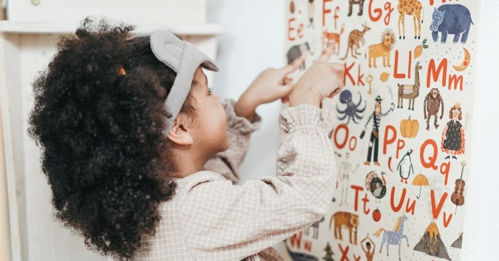 Child with curly hair points to an animal alphabet poster indoors, learning letters and animals.