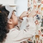 Child with curly hair points to an animal alphabet poster indoors, learning letters and animals.