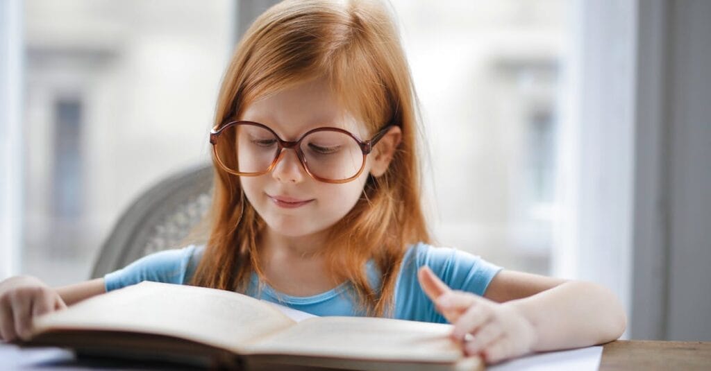 Charming young girl wearing oversized glasses reading a book at home.