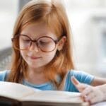 Charming young girl wearing oversized glasses reading a book at home.