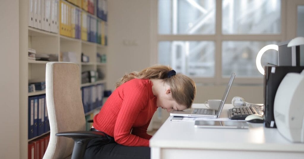 Tired woman in red sweater naps on office desk beside laptop, overwhelmed by remote work.