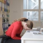 Tired woman in red sweater naps on office desk beside laptop, overwhelmed by remote work.