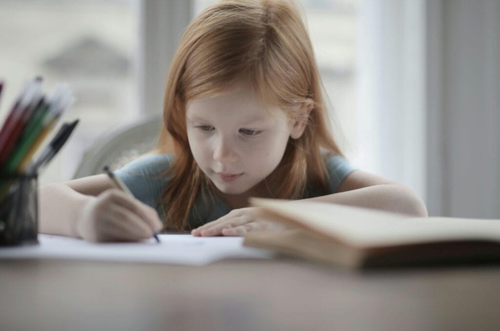 A red-haired child focused on writing at a desk with school supplies.