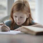 A red-haired child focused on writing at a desk with school supplies.