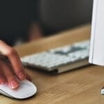 A hand using a wireless mouse at a modern desk setup with a computer and keyboard.