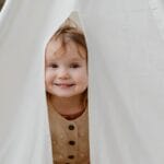 Smiling child peeks out from a cozy tent, exuding joy and playfulness in a natural setting.