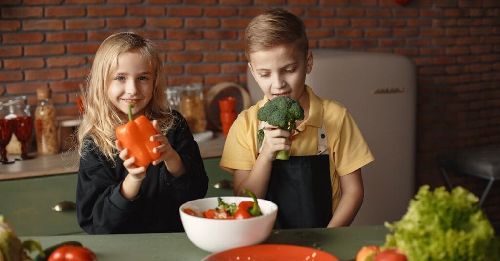 Cute girl and boy with fresh pepper and broccoli in hands standing near desk with assorted vegetables and salad bowl during vegetarian meal preparation