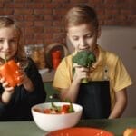 Cute girl and boy with fresh pepper and broccoli in hands standing near desk with assorted vegetables and salad bowl during vegetarian meal preparation