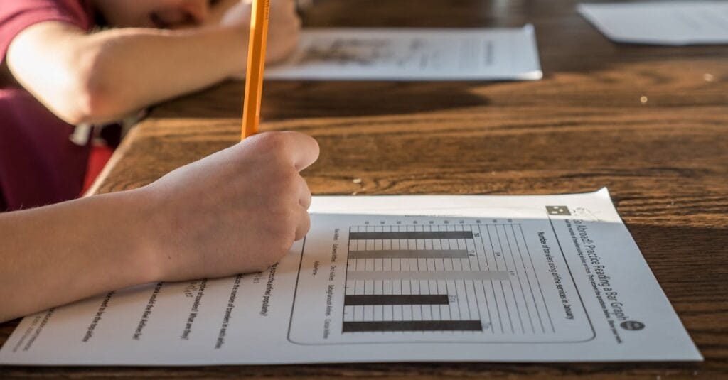 Side view of crop anonymous pupil with pencil in hand answering questions of study test with diagrams while studying at table with brother at home