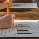 Side view of crop anonymous pupil with pencil in hand answering questions of study test with diagrams while studying at table with brother at home