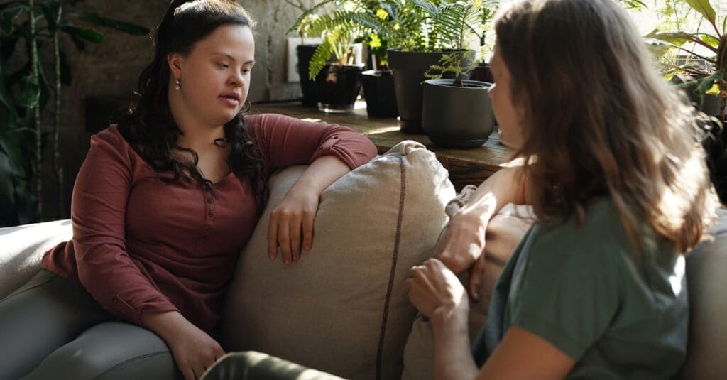 Two women having a heartfelt conversation on a cozy sofa surrounded by green plants indoors.