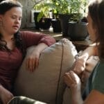 Two women having a heartfelt conversation on a cozy sofa surrounded by green plants indoors.