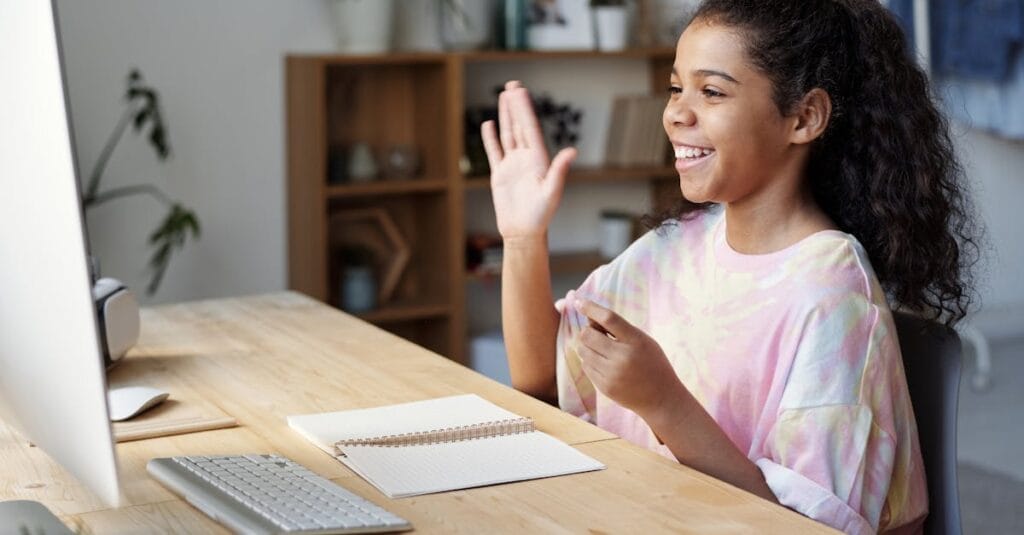 A happy girl waves during an online class at home, sitting at a desk with a computer.