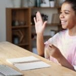 A happy girl waves during an online class at home, sitting at a desk with a computer.