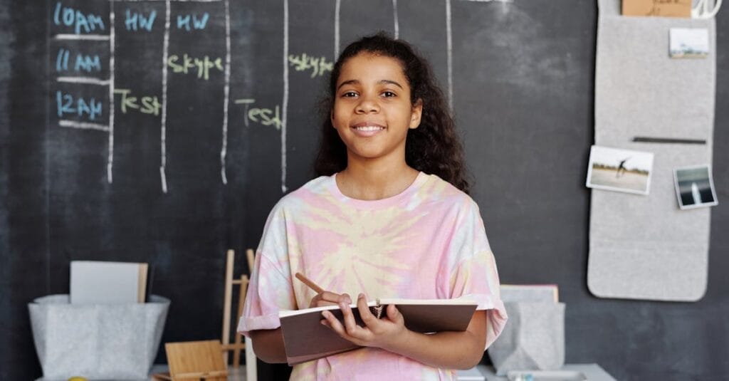 Smiling teen girl holding a notebook, standing in a classroom with a chalkboard timetable.