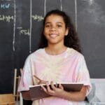 Smiling teen girl holding a notebook, standing in a classroom with a chalkboard timetable.