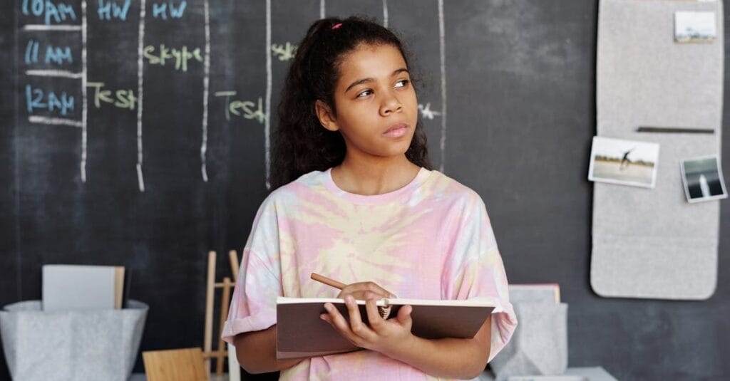 A teenager in a pink shirt ponders while holding a notebook in a classroom setting.