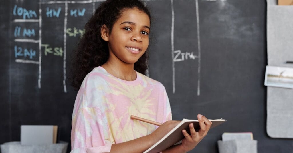 Teenage girl smiling while holding a notebook in a classroom setting with a chalkboard behind her.