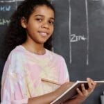 Teenage girl smiling while holding a notebook in a classroom setting with a chalkboard behind her.