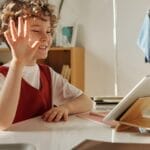 Happy child waving during an online learning session at home.