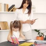 Smart little girl sitting at table with abacus and textbooks and writing in diary with pencil with content young home teacher in white shirt standing behind during early home education