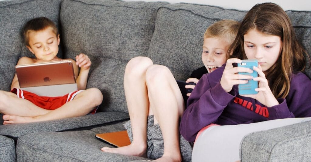 Positive barefoot children in casual wear resting together on cozy couch and browsing tablets and smartphones