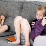 Positive barefoot children in casual wear resting together on cozy couch and browsing tablets and smartphones