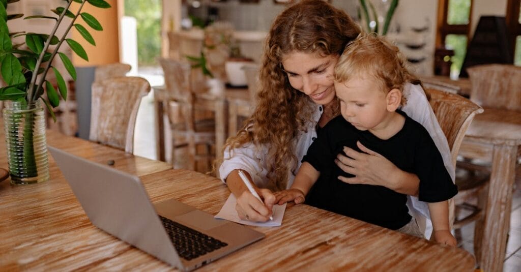 A mother assists her young son in writing while using a laptop at a wooden table.