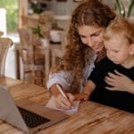 A mother assists her young son in writing while using a laptop at a wooden table.