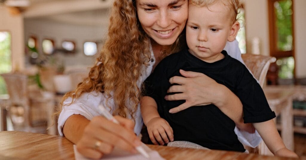 Mother holding her young child while writing, creating a bonding moment indoors.